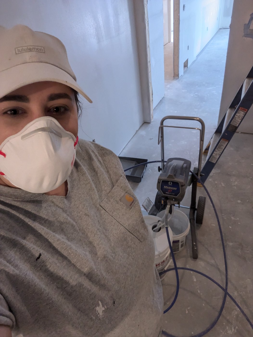 Cyan Goulet in a dust mask beside her airless paint sprayer in a hallway being painted